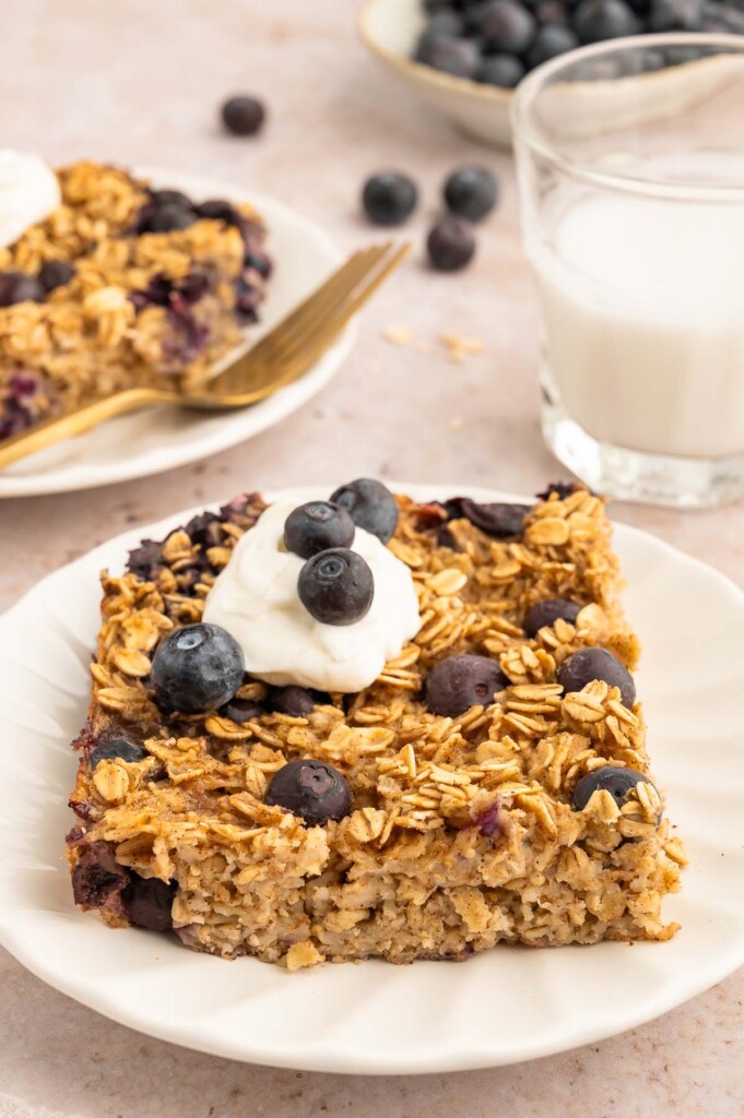 A white plate of blueberry baked oatmeal topped with whipped cream and blueberries with another plate in the background.