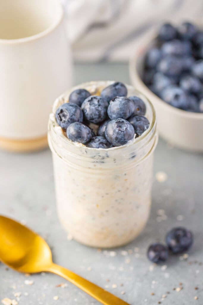 Overnight blueberry oats in a jar with berries and a gold spoon on the side on a grey background with a bowl of blueberries in the back