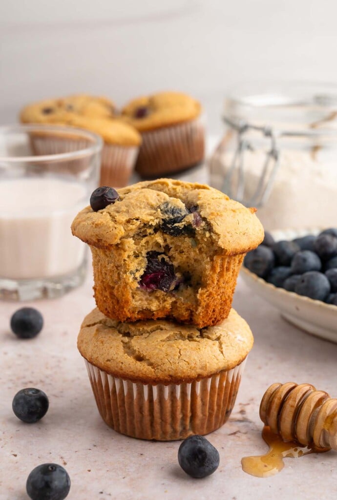 A blueberry protein muffin with a bite taken out of it on top of another muffin with more muffins in the background with blueberries on the side. 