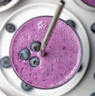 A close up of a blueberry smoothie in a clear glass with a straw sticking out of it