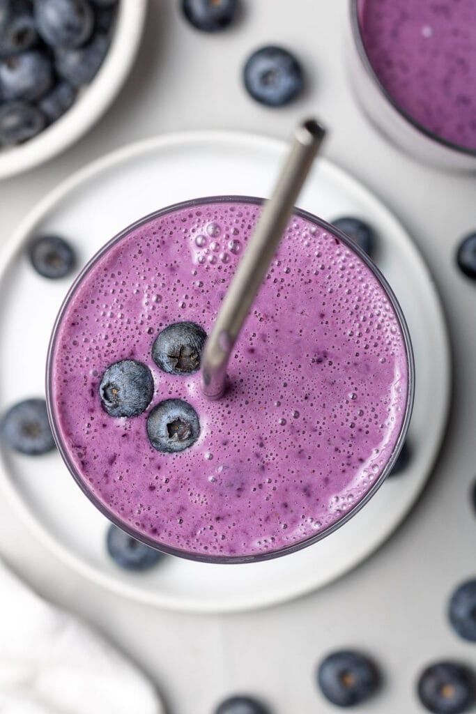 A close up of a blueberry smoothie in a clear glass with a straw sticking out of it