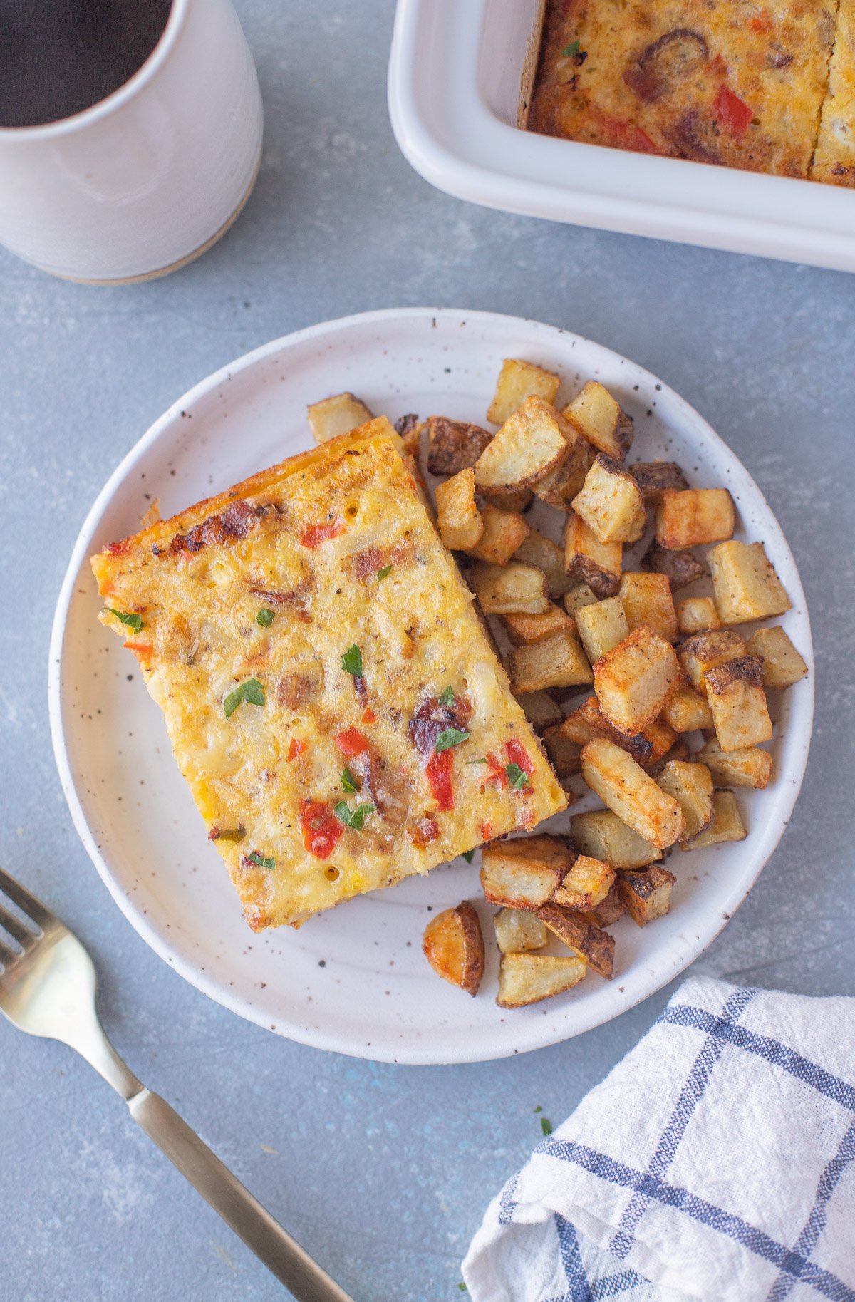 An overhead shot of a white plate with a piece of bacon breakfast casserole and roasted potatoes with another casserole dish, a mug of coffee, and fork on the side