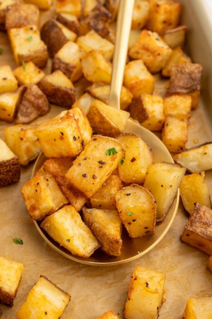 Breakfast potatoes in a large gold spoon on a parchment lined baking sheet with more potatoes.