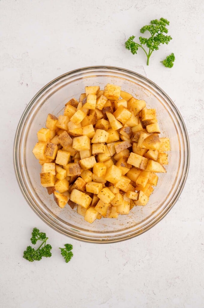 Raw chopped russet potatoes in a clear bowl tosses with olive oil and spices. 