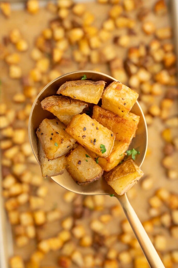 A gold spoon holding up breakfast potatoes with more potatoes on a baking sheet in the background.