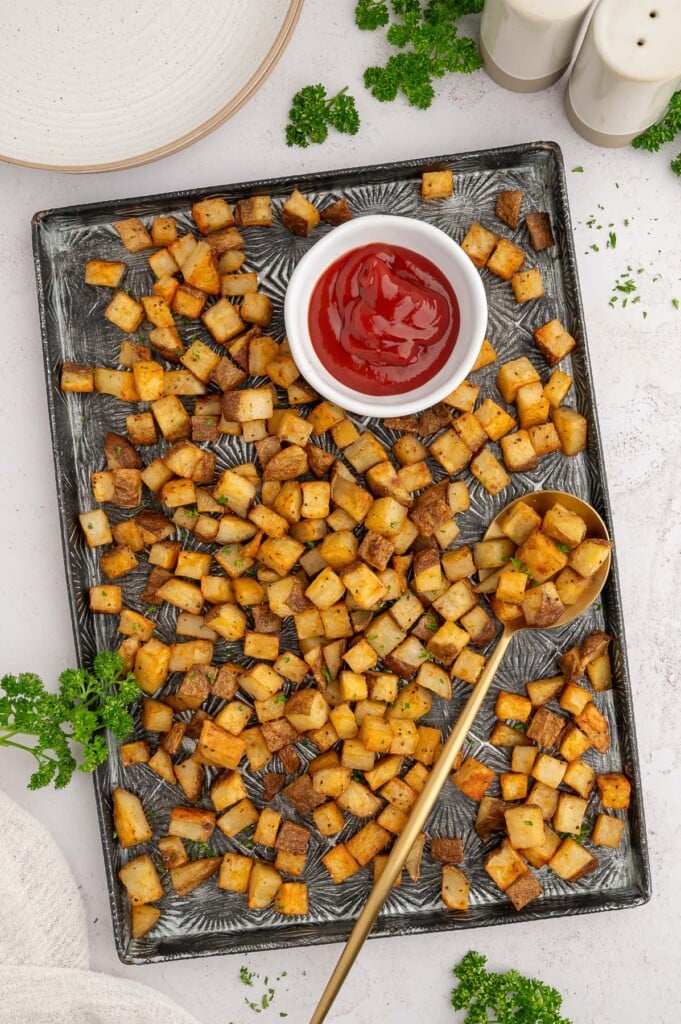 Breakfast potatoes on a black baking sheet with a spoon and a white bowl of ketchup on the sheet.