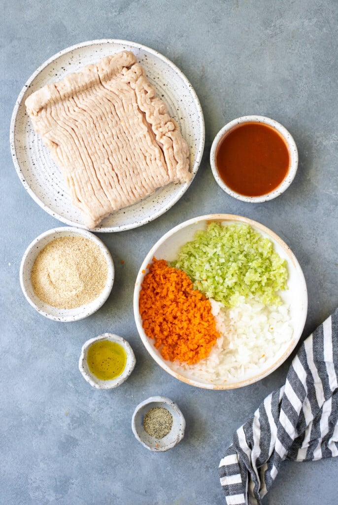 Ingredients for buffalo chicken meatballs in white bowls: ground chicken, carrots, celery, onions, olive oil, buffalo sauce, pepper, bread crumbs, blue cheese 