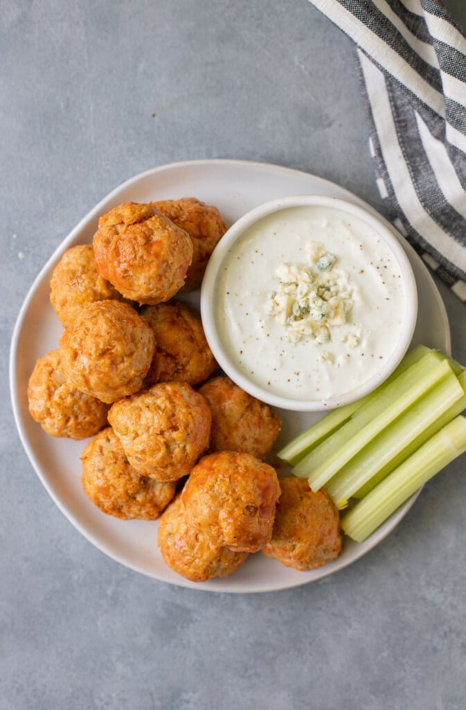 An overhead shot of buffalo chicken meatballs on a white plate with a bowl of blue cheese and celery on a grey background