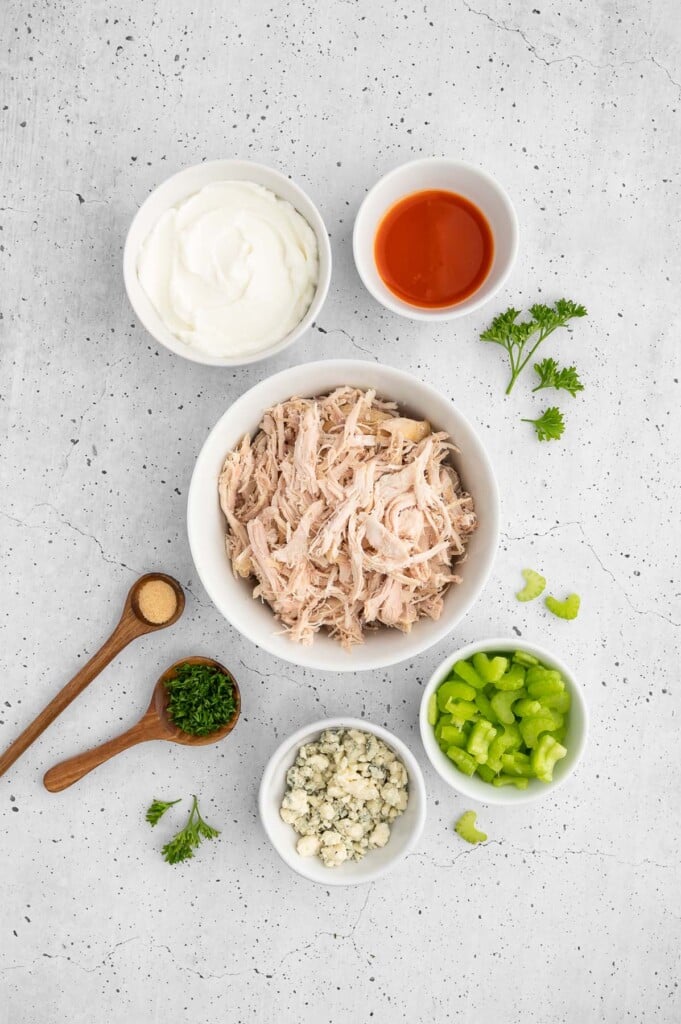 Ingredients for buffalo chicken salad in small bowls on a grey background: cooked shredded chicken, nonfat greek yogurt, celery, fresh parsley, garlic powder, buffalo sauce, blue cheese crumbles