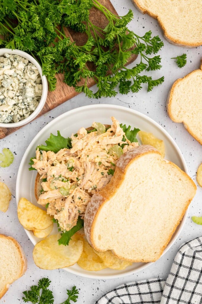 buffalo chicken salad on top of lettuce with a piece of bread slightly to the side on a white plate with chips on the side on a grey background. 