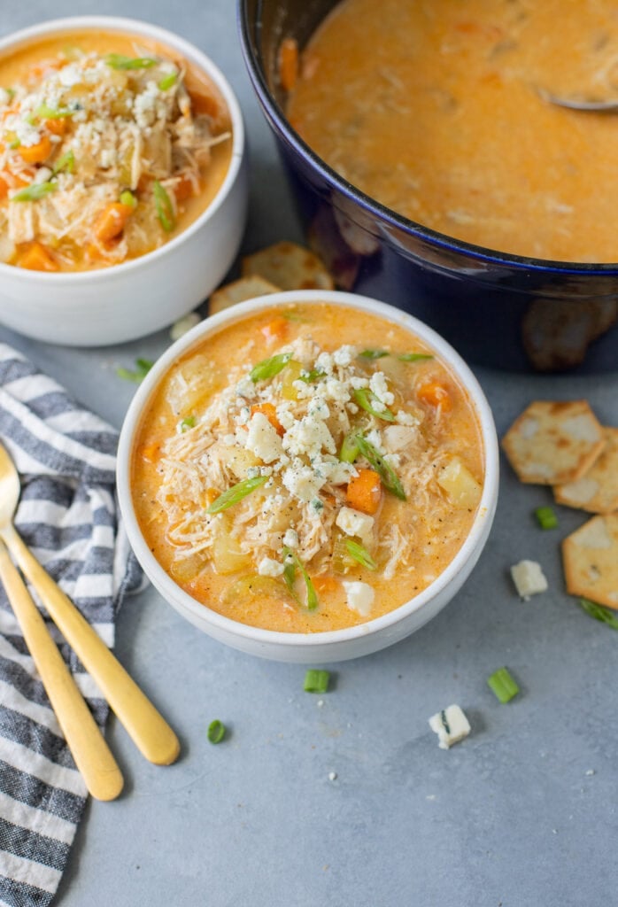 Two bowls of buffalo chicken soup beside a pot of buffalo chicken soup on a grey background with spoons on the side