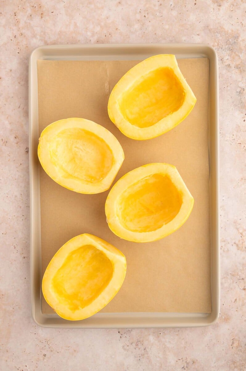 Four prepped spaghetti squash halves on a parchment lined baking sheet. 