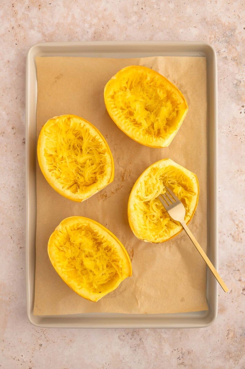 Cooked spaghetti squash boats after shredding with a fork left in one. All are on a parchment lined baking sheet.