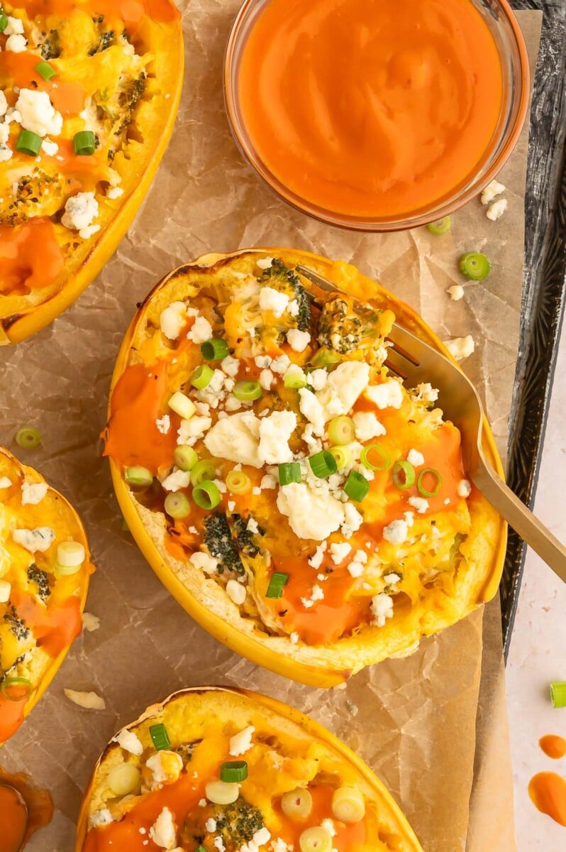 Buffalo chicken spaghetti squash boat with a fork in it and more squashes and a bowl of buffalo sauce on the side.