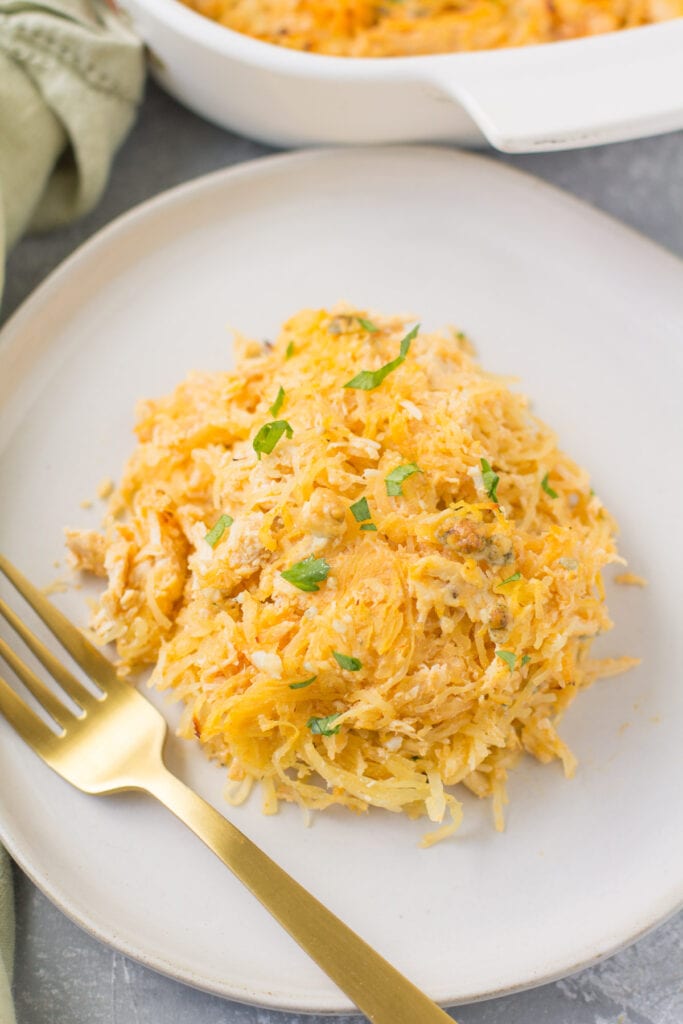 A white plate with a scoop of healthy buffalo chicken casserole on it. a gold fork is off to the left with a casserole dish in the background