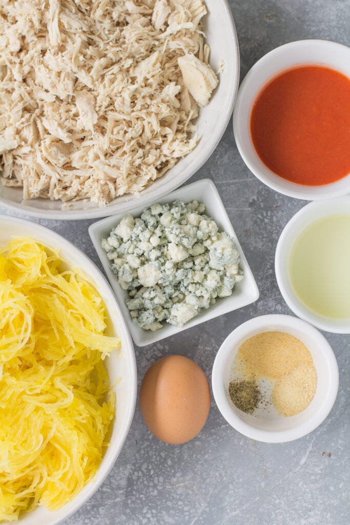 Ingredients for Buffalo Chicken Spaghetti Squash Casserole on a grey background all laid out in small white bowls.