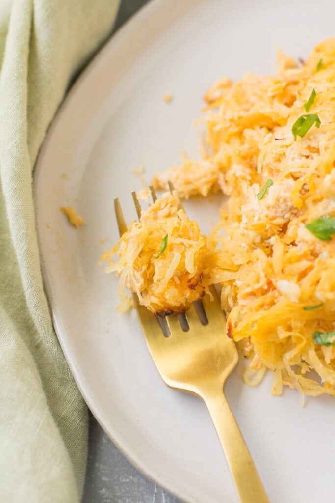 A gold fork taking a bite out of buffalo chicken spaghetti squash casserole on a white plate.