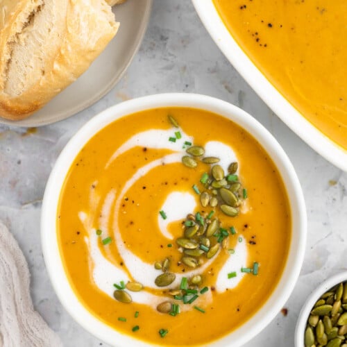 An overhead shot of a white bowl of instant pot butternut squash soup with cream, herbs and pumpkin seeds on top with a dish of soup and bread on the side