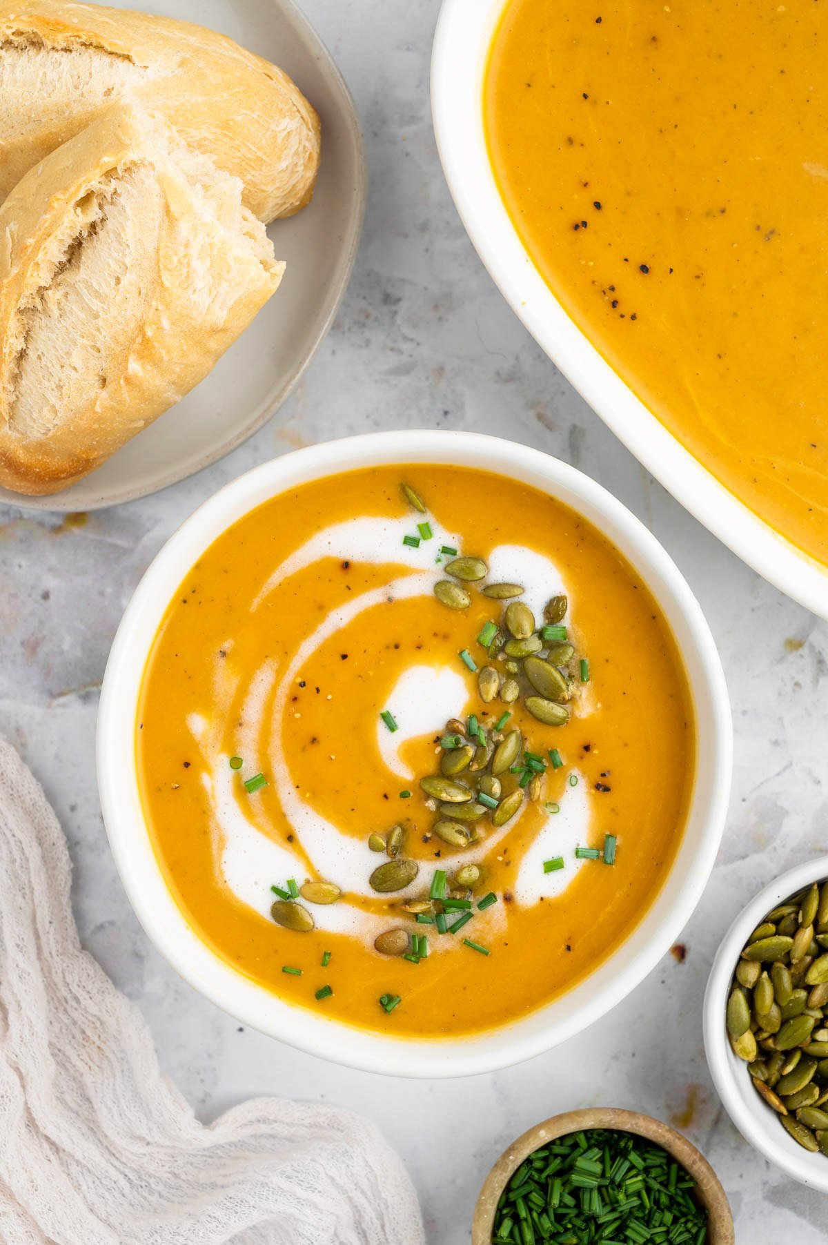 An overhead shot of a white bowl of instant pot butternut squash soup with cream, herbs and pumpkin seeds on top with a dish of soup and bread on the side