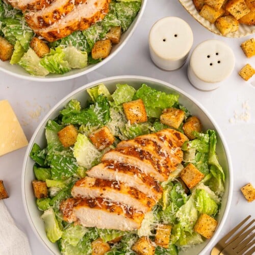 An overhead shot of a grilled chicken caesar salad topped with homemade croutons in a bowl with another salad on the side.