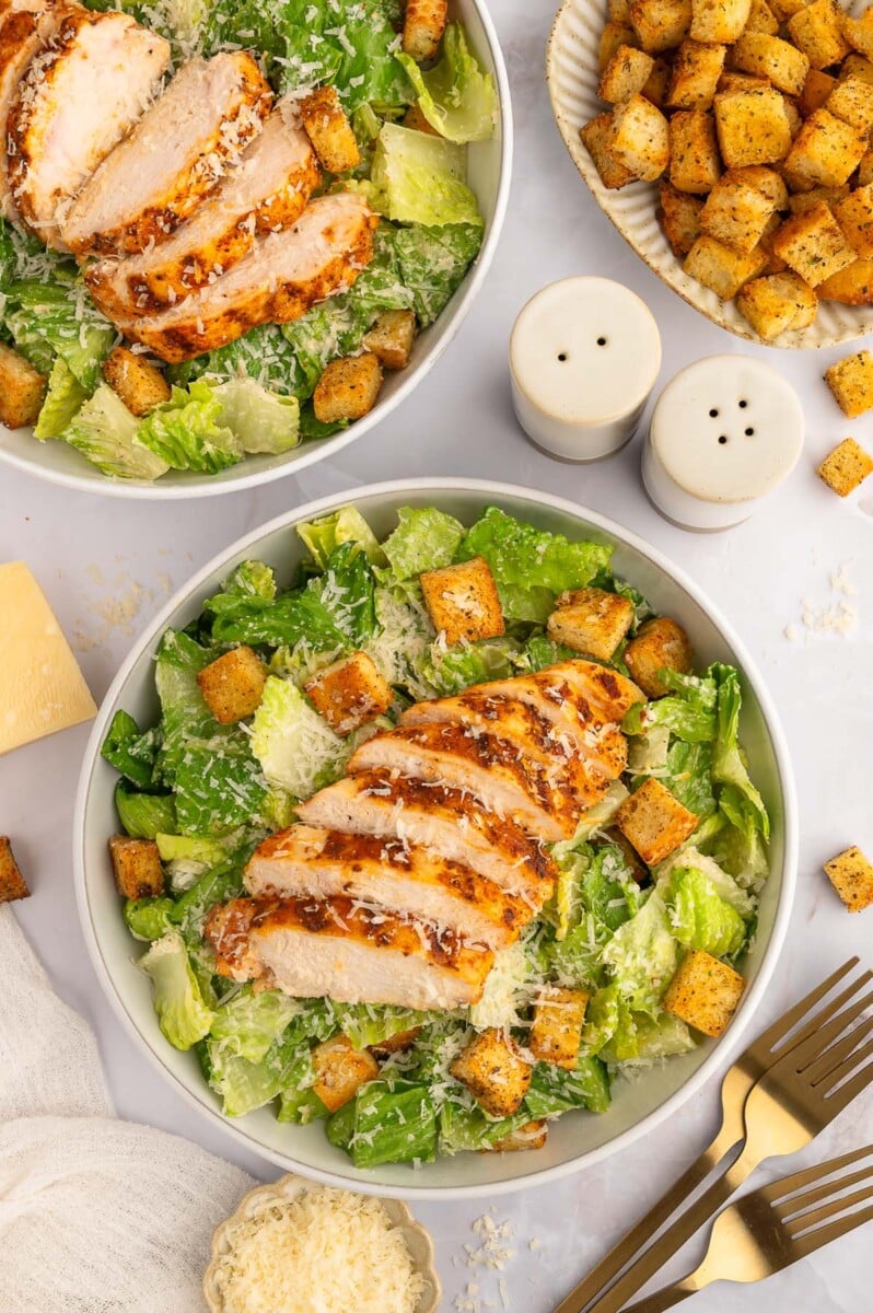 An overhead shot of a grilled chicken caesar salad topped with homemade croutons in a bowl with another salad on the side.