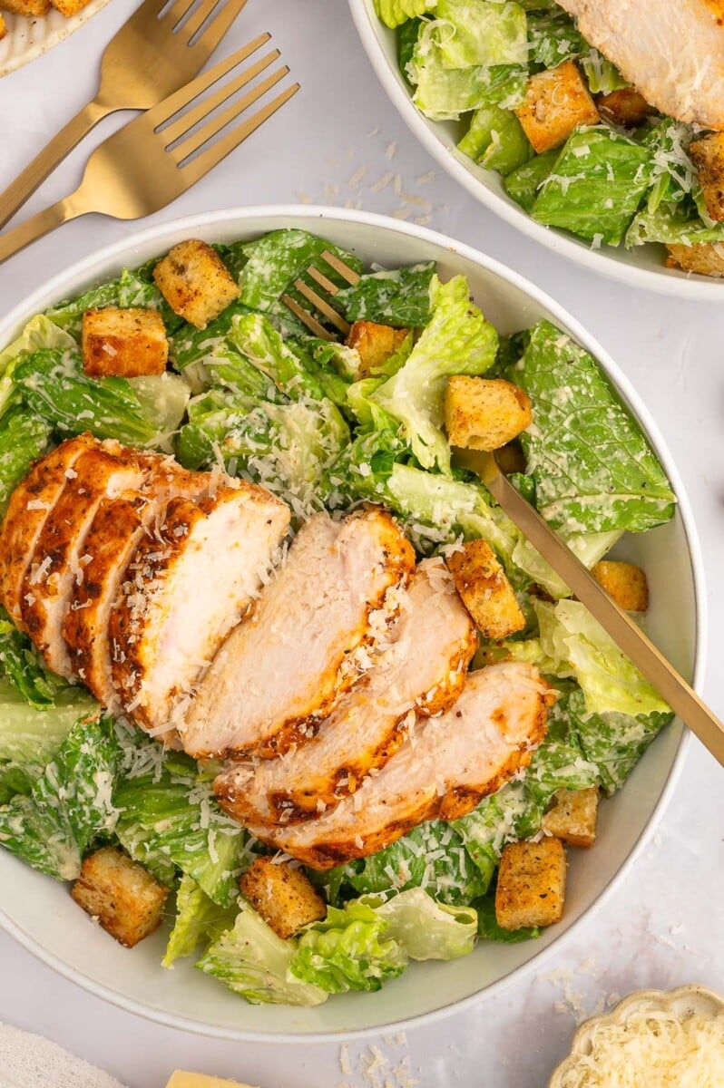 A sliced grilled chicken caesar salad in a white bowl topped with homemade croutons and a fork in the bowl.