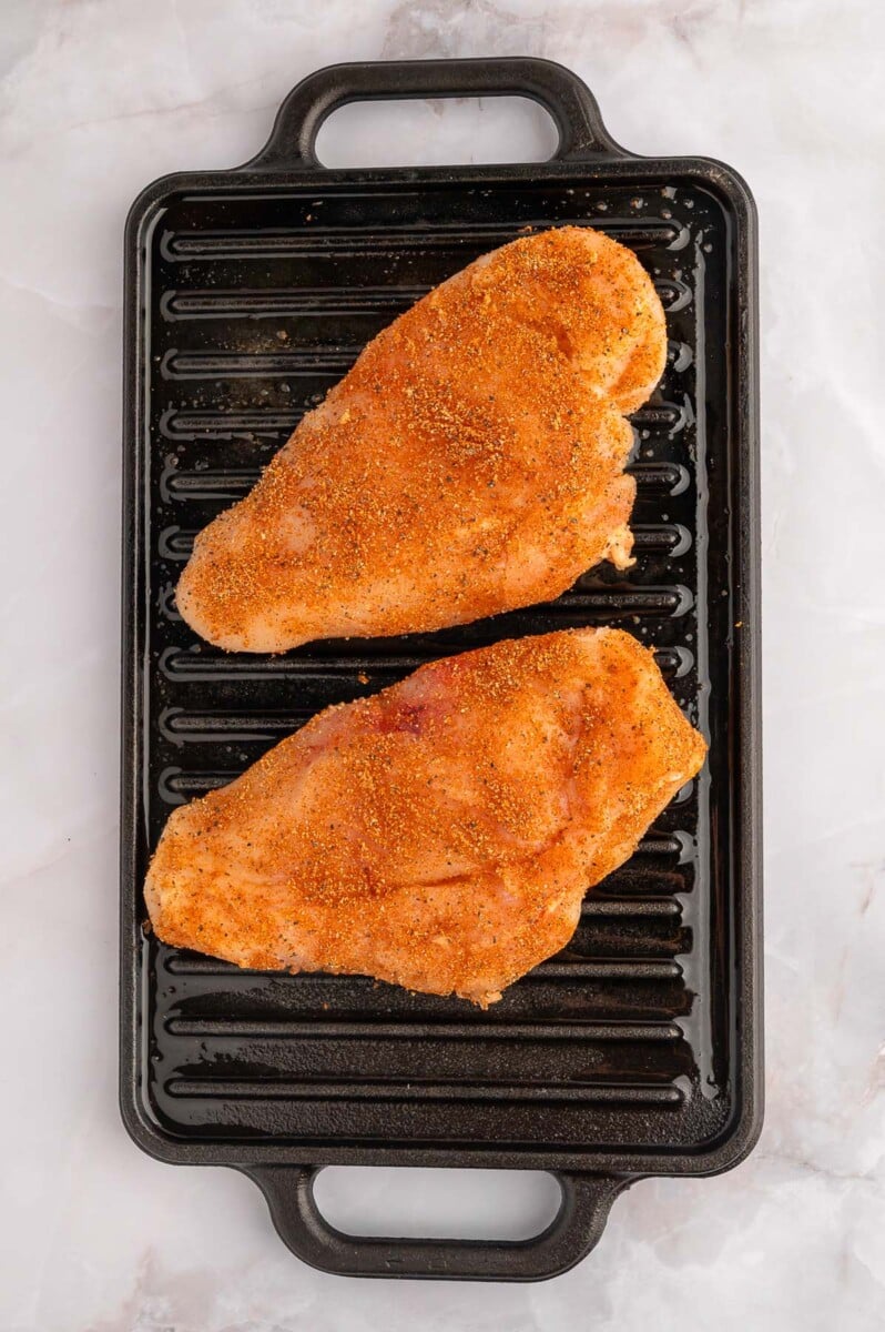 Two seasoned raw chicken breasts on a black stovetop grill.