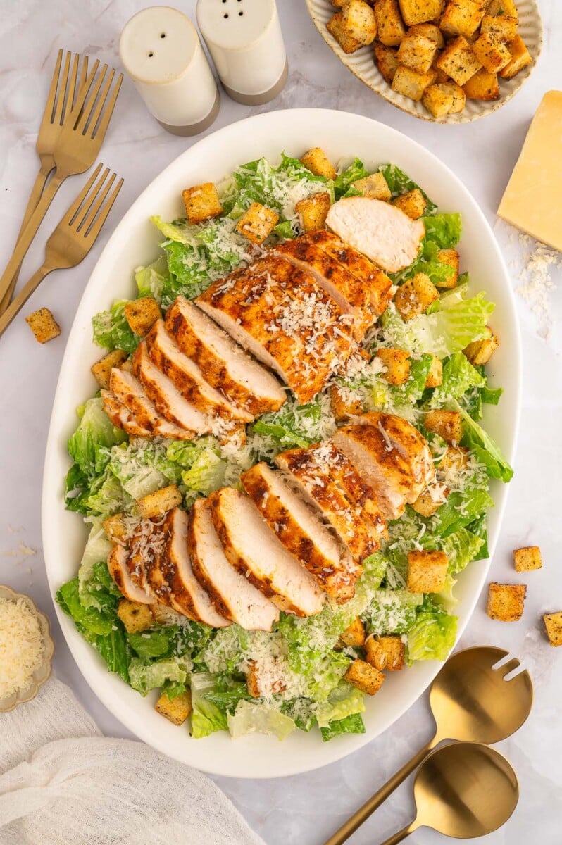 A white platter of a caesar salad with two sliced grilled chicken breasts on top. 