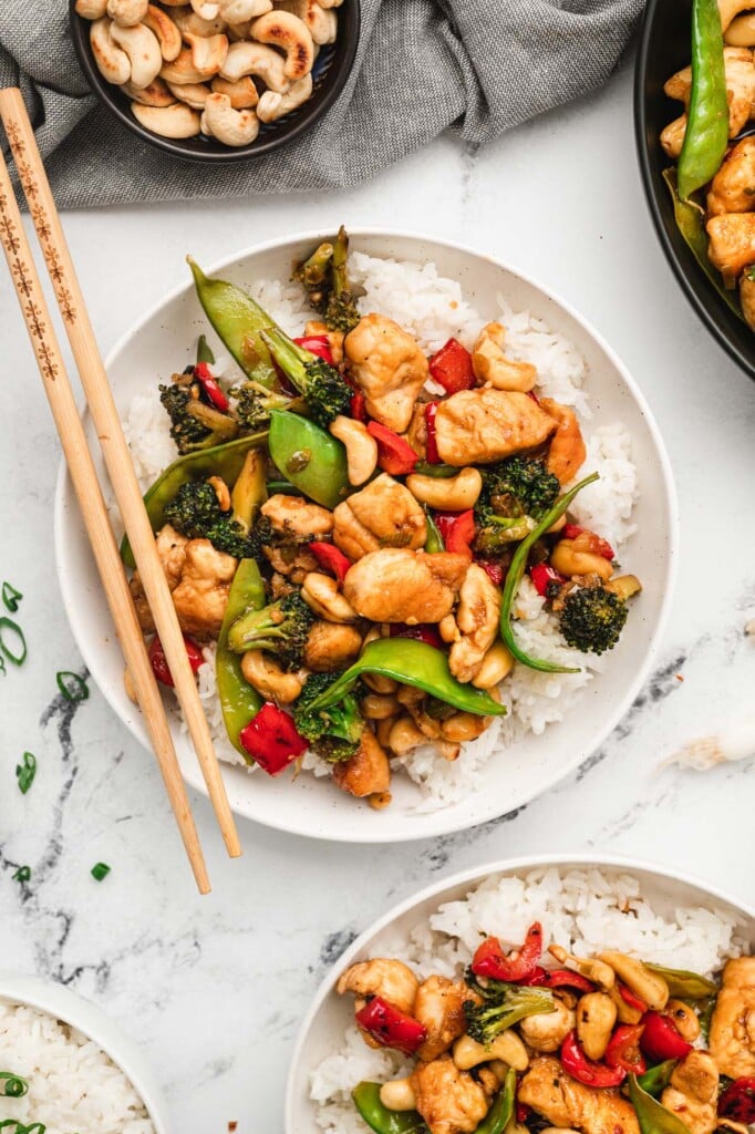 An overhead shot of a white plate with cashew chicken on a bed of white rice with chopsticks, another plate and a pan of cashew chicken on the side
