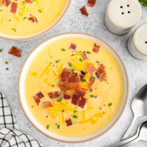 An overhead shot of a bowl of cauliflower soup topped with cheese and bacon with spoons, a towel and another bowl of soup on the side