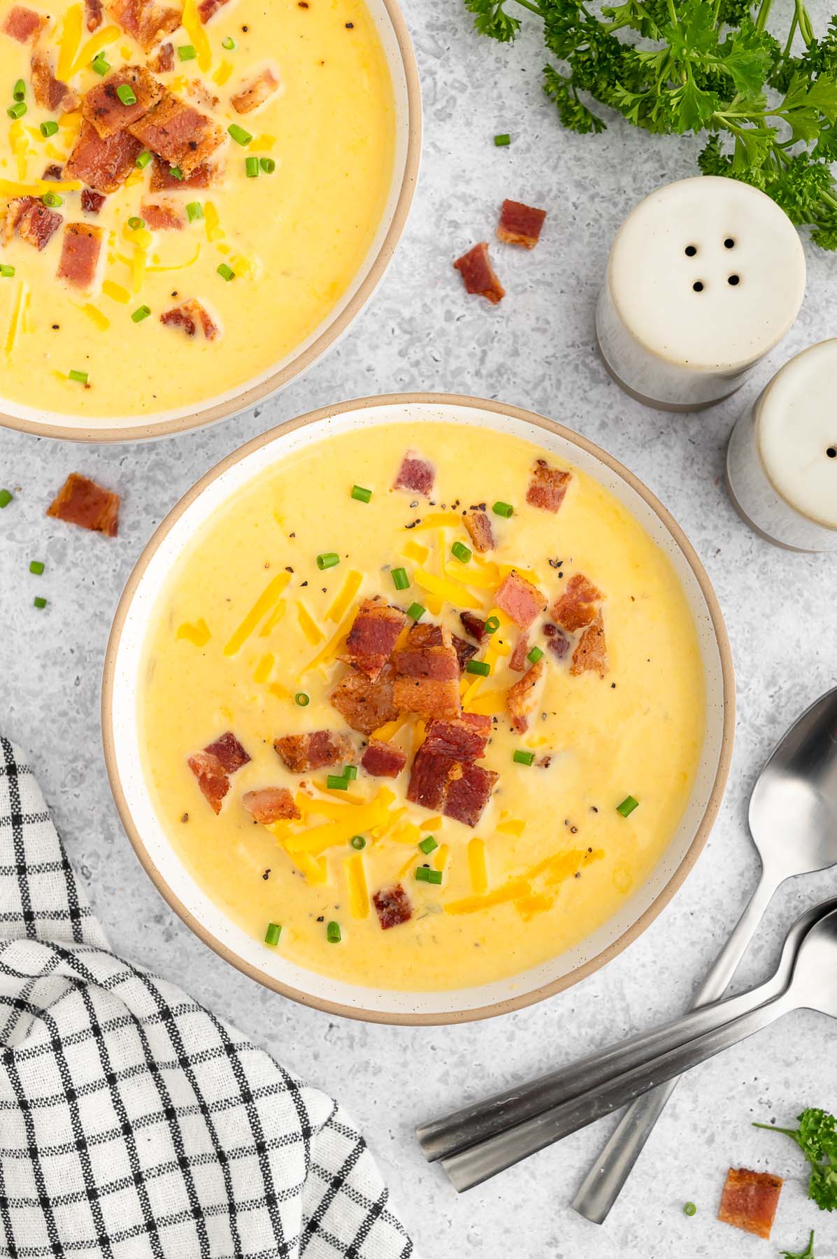An overhead shot of a bowl of cauliflower soup topped with cheese and bacon with spoons, a towel and another bowl of soup on the side