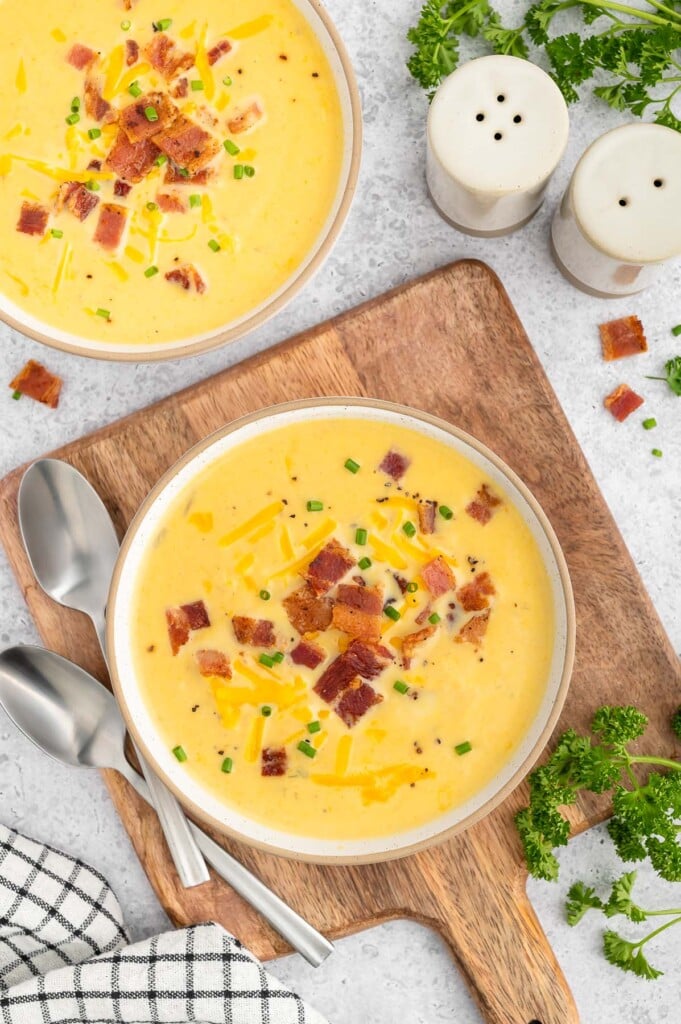 An overhead shot of a bowl of cauliflower soup topped with cheese and bacon with spoons, a towel and another bowl of soup on the side