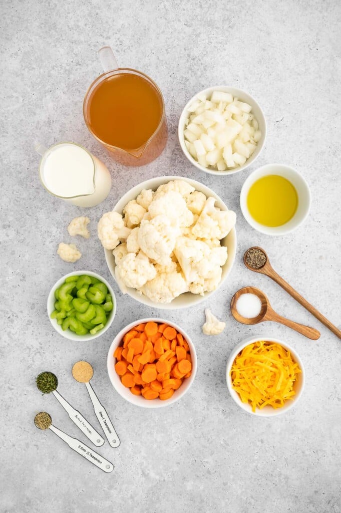 Ingredients for cauliflower soup recipe in white bowls: olive oil, onions, carrots, celery, cauliflower florets, vegetable broth, garlic powder, dried parsley, dried rosemary, salt, pepper, milk of choice, cheddar cheese