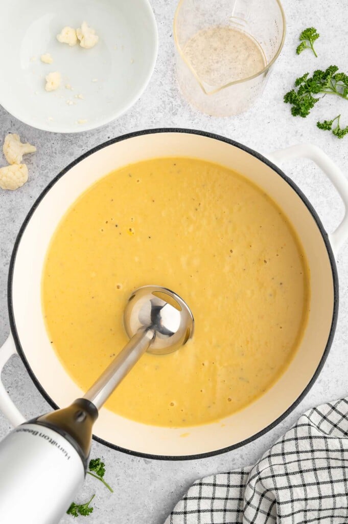An overhead shot of a stock pot of creamy cauliflower soup after cooking while an immersion blender is blending