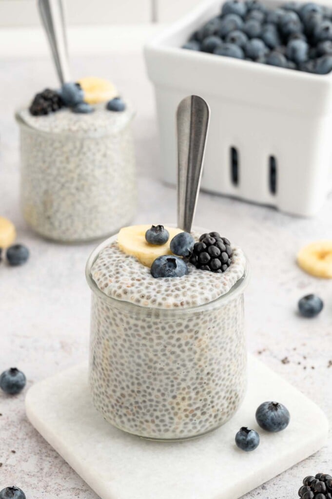 Vanilla Chia Seed Pudding in a clear jar topped with blueberries, blackberries, and sliced banana with a spoon in the jar and a white container of blueberries in the background.