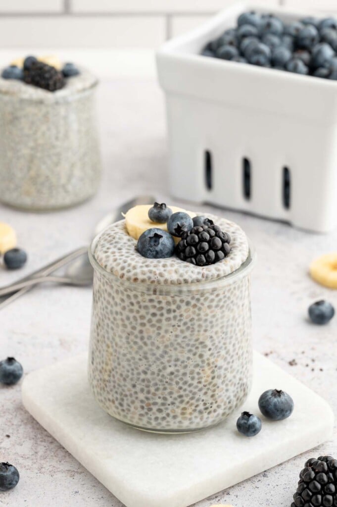 Vanilla Chia Seed Pudding in a clear jar topped with blueberries, blackberries, and sliced banana with a white container of blueberries in the background.