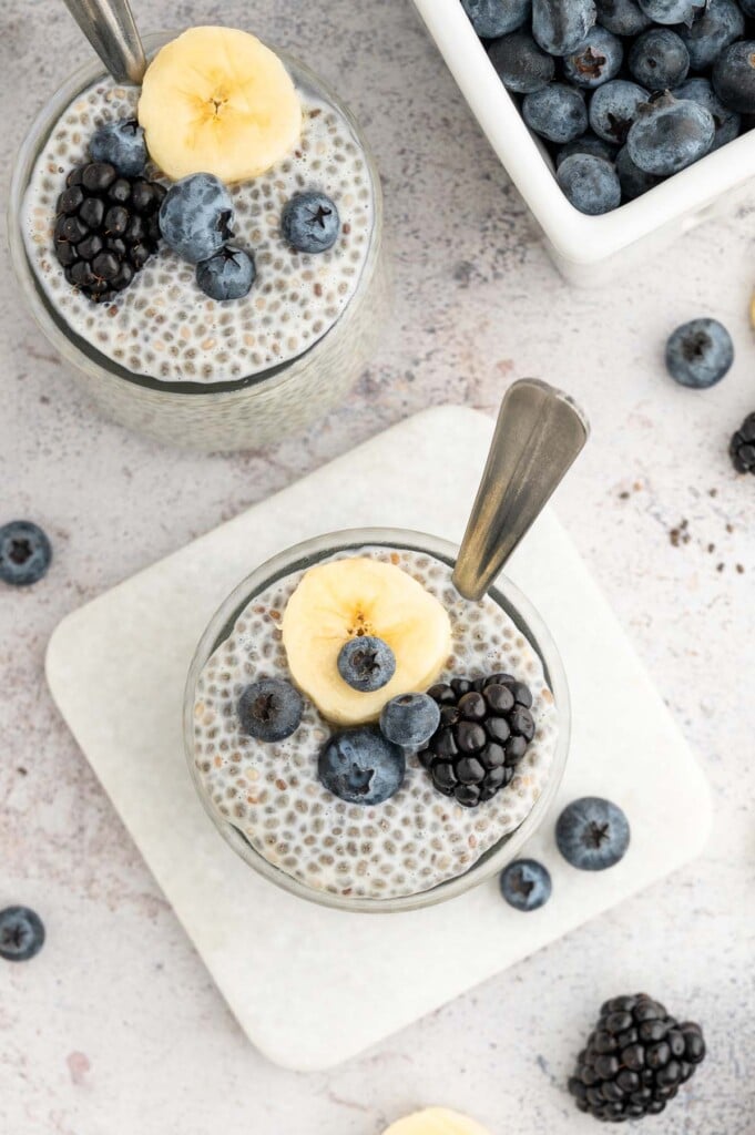 An overhead shot of two jars of chia seed pudding topped with berries and banana slices with spoons in it.