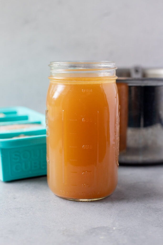 A close up of a container of homemade chicken broth with a pot off to the side