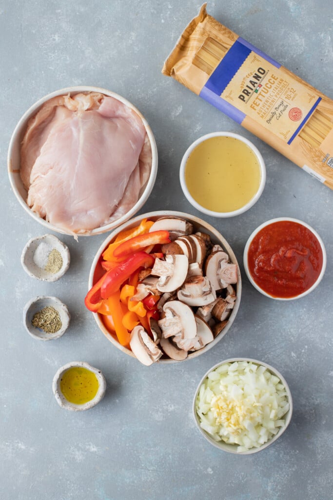 Ingredients for chicken cacciatore in white bowls: Olive oil, Chicken breasts, Salt, Pepper, Onion, Garlic, Mushrooms, Bell peppers, Red wine, Crushed tomatoes, Chicken broth, Thyme, Rosemary , Oregano, Pasta