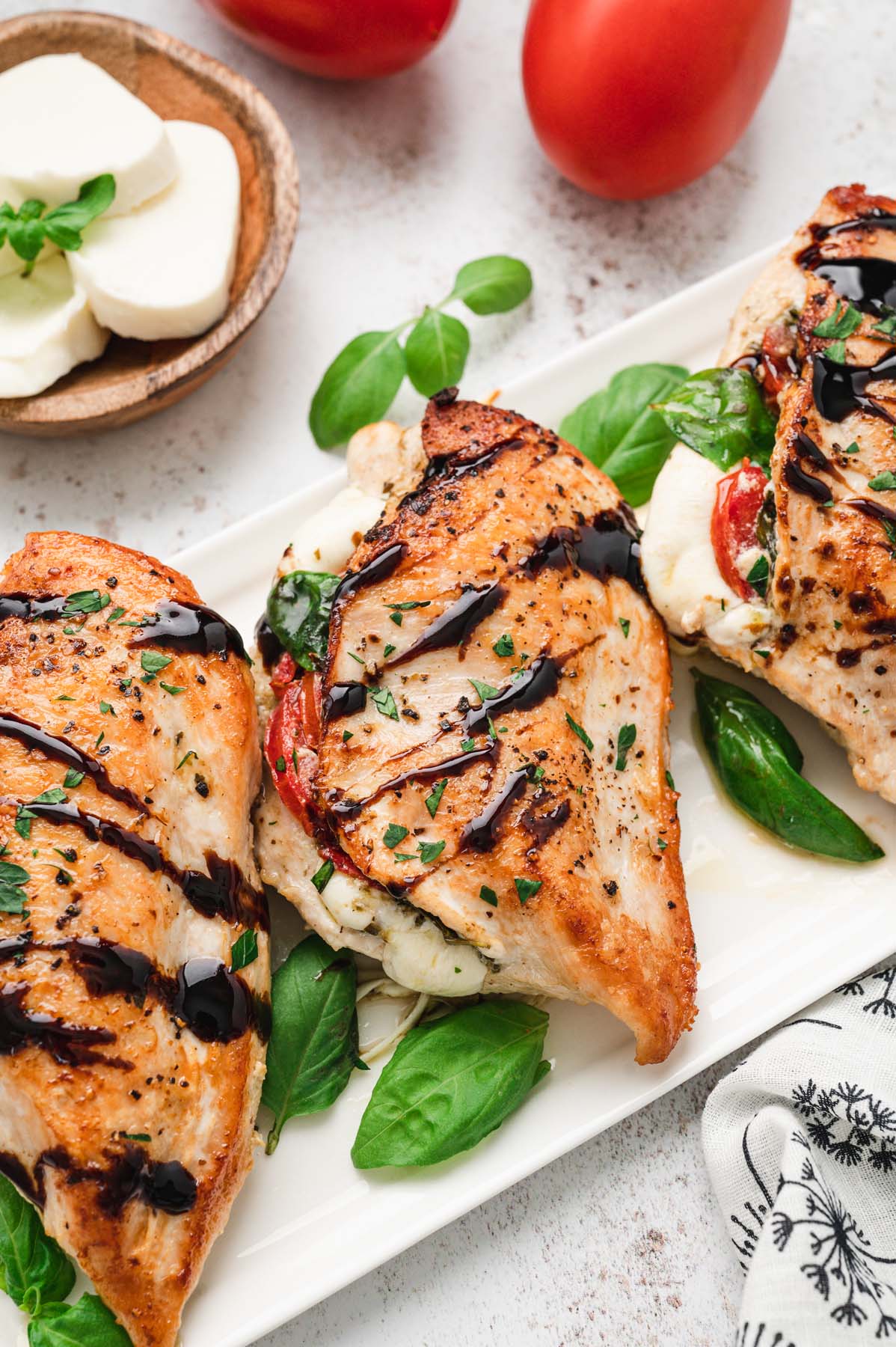 Three chicken caprese breasts on a white platter topped with balsamic glaze and fresh basil leaves with mozzarella and tomatoes on the side