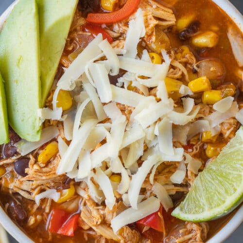 A close up of Chicken Enchilada Soup in a white bowl with avocado, lime and cheese on top
