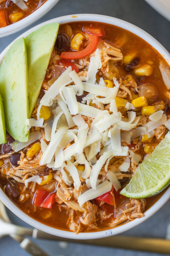 A close up of Chicken Enchilada Soup in a white bowl with avocado, lime and cheese on top
