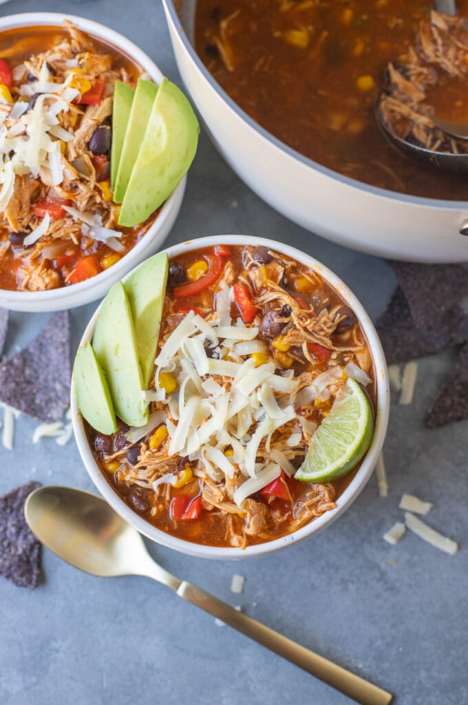 a bowl of cheesy chicken enchilada soup on a grey background with a separate bowl and bot off to the side.