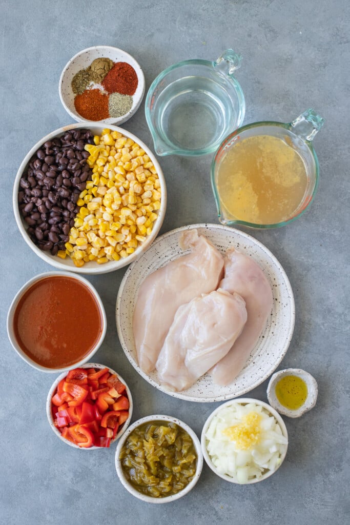 Ingredients for Enchilada Soup in small white bowls on a grey background: olive oil, onion, garlic, bell peppers, boneless skinless chicken breast, homemade enchilada sauce, diced green chiles, chicken broth, paprika, pepper, ground cumin, chili powder, oregano, black beans, frozen corn