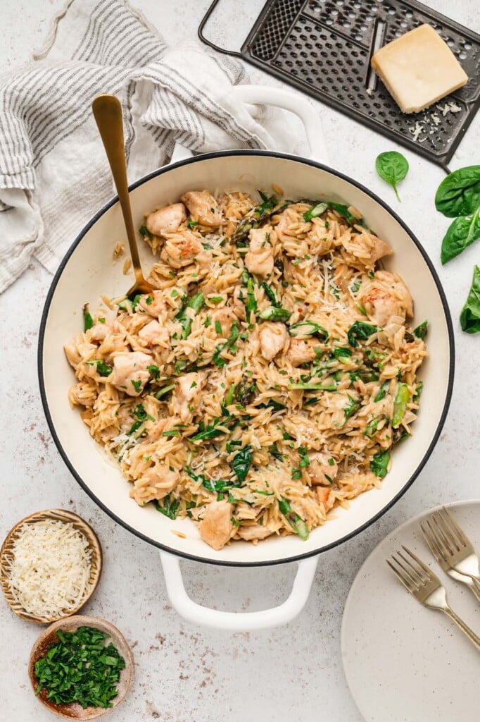 An overhead shot of chicken orzo in a dutch oven with parsley and parmesan on the side in small bowls