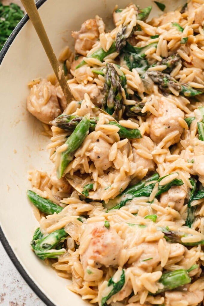 A close up of chicken orzo in a cream dutch oven with a gold spoon taking a scoop out of it