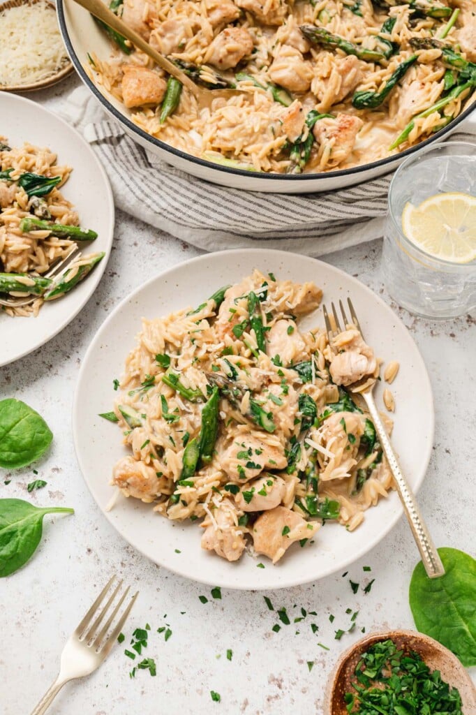 An overhead shot of a serving of chicken orzo on a white plate with another plate and dutch oven on the side