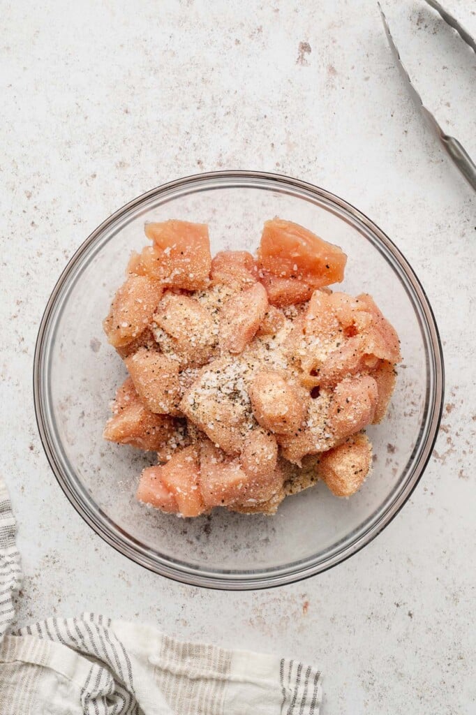 An overhead shot of chicken chunks, garlic powder, salt, pepper and red pepper flakes in a clear bowl on a counter