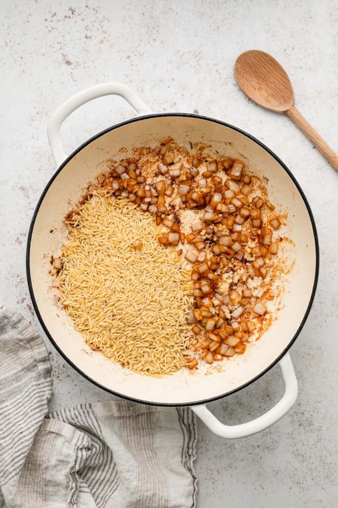 Softened onions, uncooked orzo and garlic in a cream dutch oven with a wooden spoon on the side