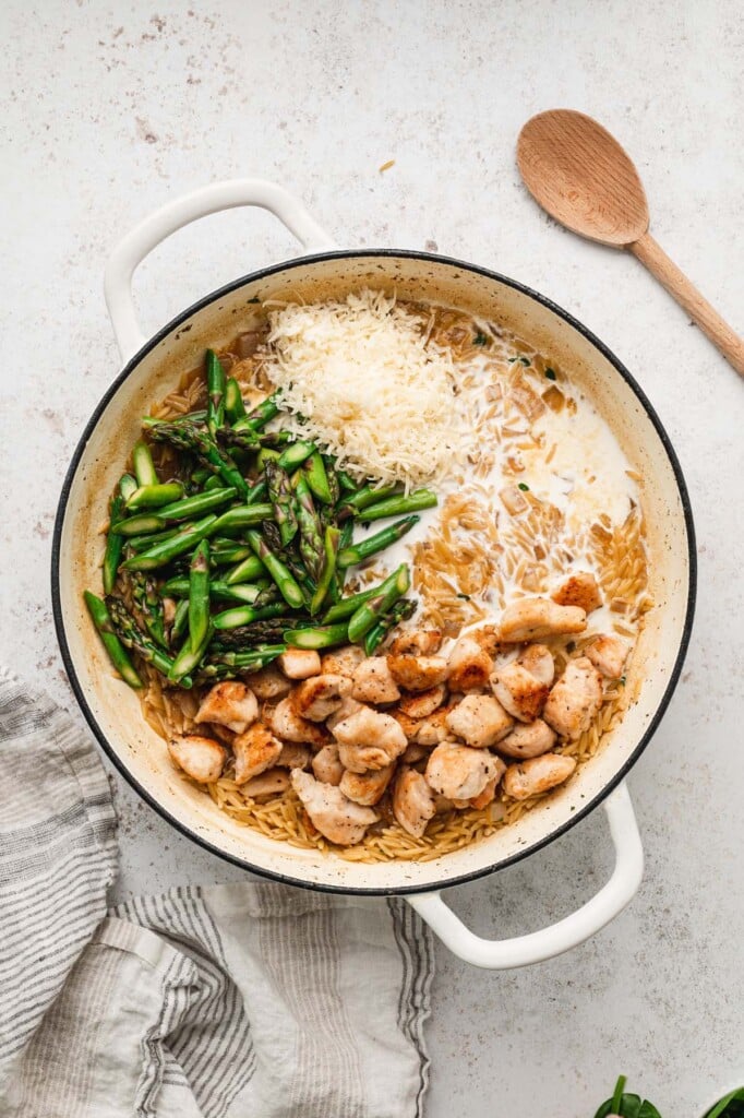 An overhead shot of browned chicken, half and half, asparagus, parmesan being added to a dutch oven of orzo before being mixed in with a wooden spoon on the side